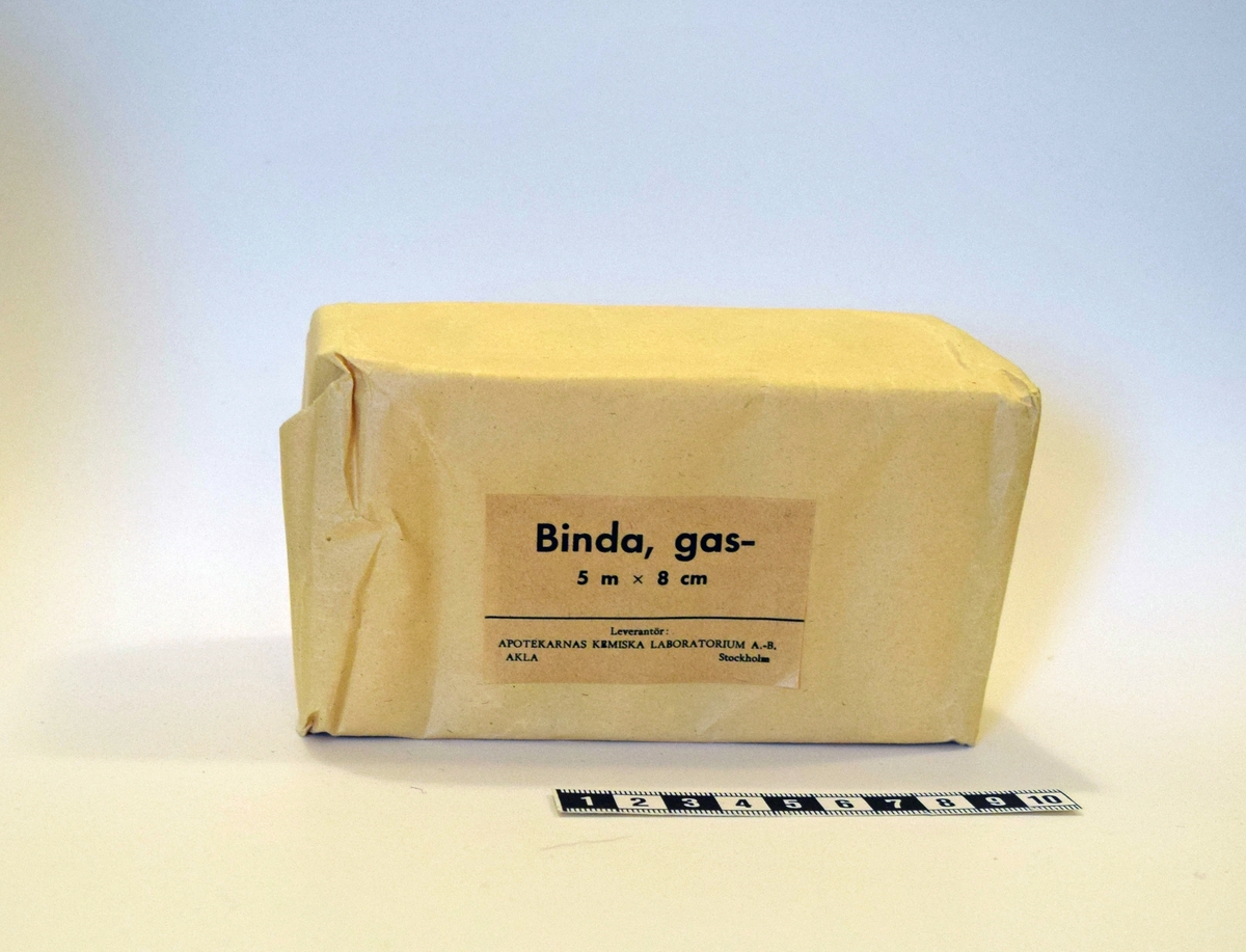 Tio rullar med gasbinda som förvaras i förpackning av gult papper. Tejpad med papperstejp. Etikett på framsidan.