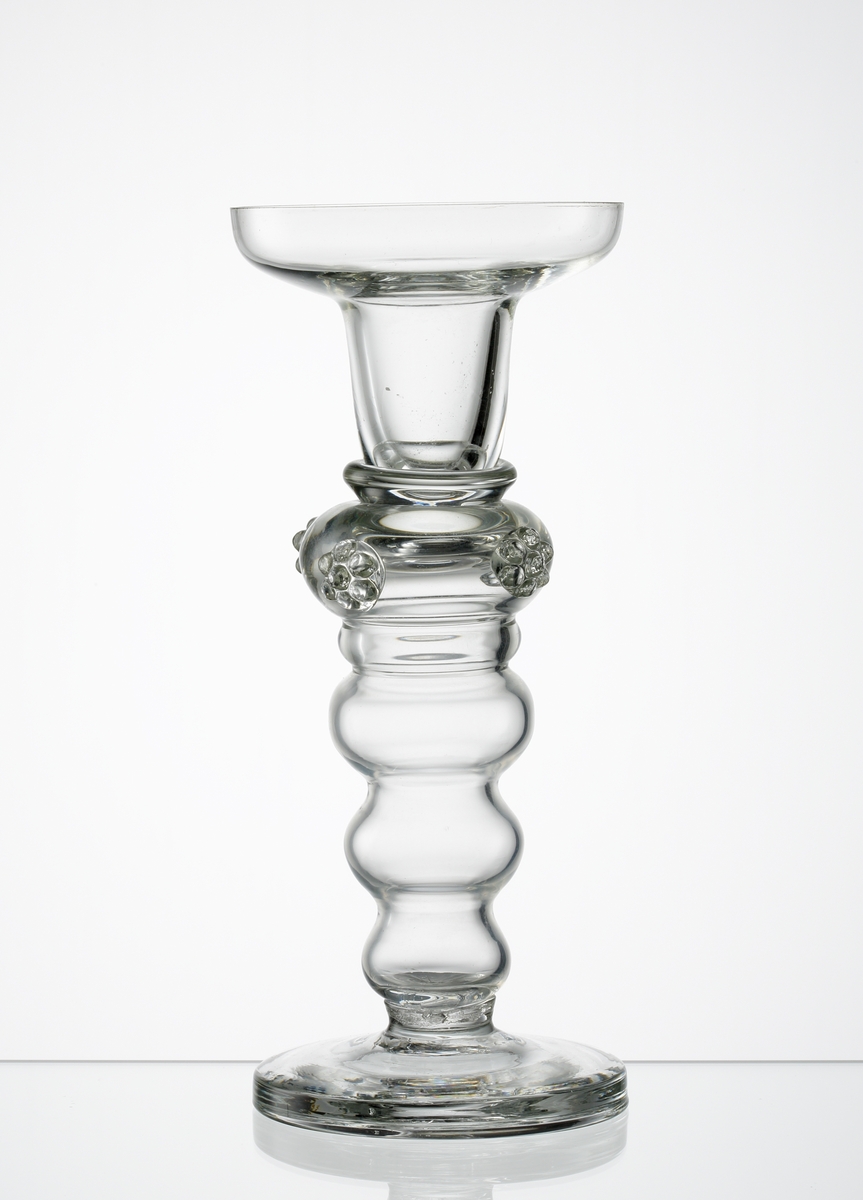 Ljusstake i ofärgat glas, med ben bestående av fyra vulster, varav den översta under ljuspipan försetts med fyra "hallon". Ljuspipa med manschett blåst i ett stycke.