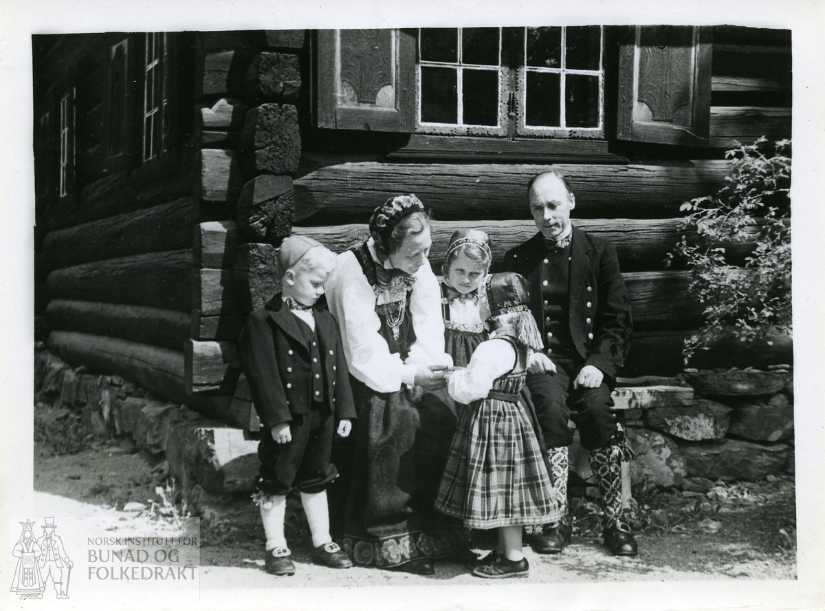 Kvinne, mann og barn i bunader foran tømmerhus.