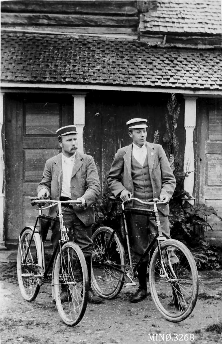 To menn med hver sin sykkel mot en husvegg. 