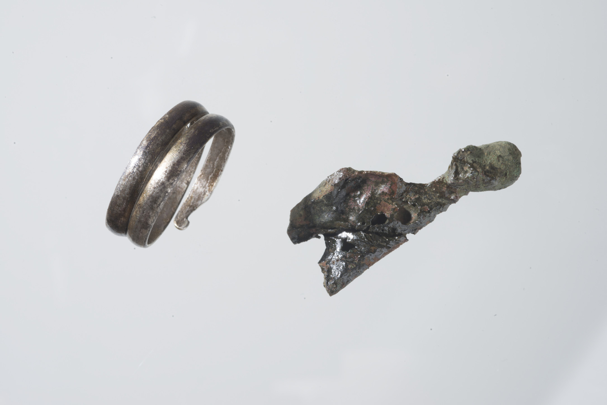 Fragment, mulig del av spenne, st.l. 37 mm, st.b. 15 mm.  Avlangt fragment med et bøyd hode i en kortende og avknekt i motsatt ende. Overflaten på den ene siden har et bevart sort korrosjonslag. Uviss alder.