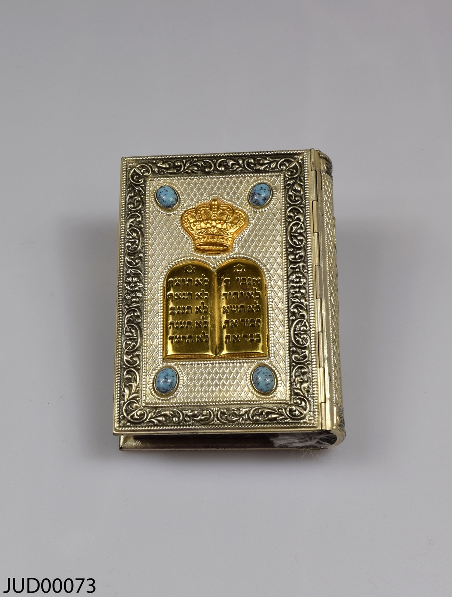 Siddur med silverpärm, liggandes i pappkartong. Bokens framsida är dekorerad med förgyllda stentavlor med krona på toppen, fyra turkosa stenar i varsitt hörn. Boken är skriven på hebreiska och tryckt i Israel 1960.