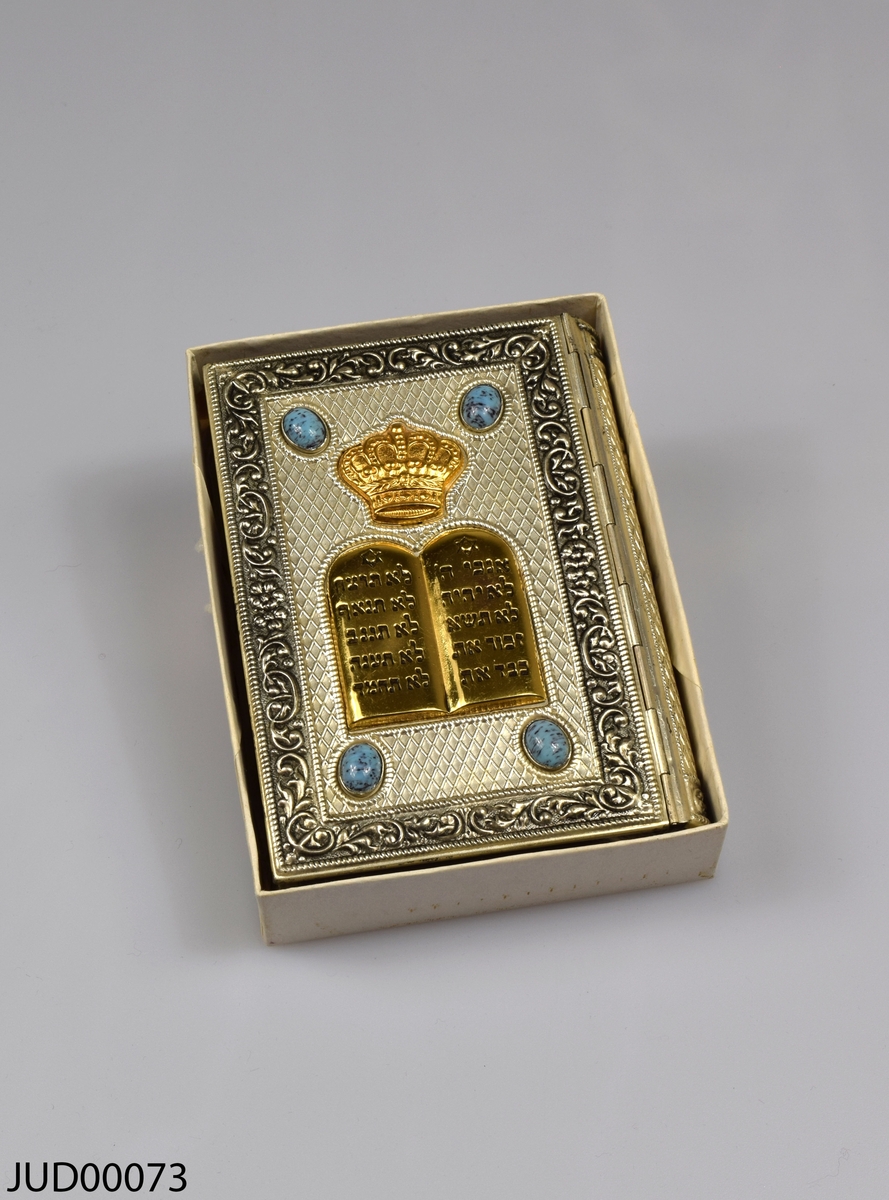 Siddur med silverpärm, liggandes i pappkartong. Bokens framsida är dekorerad med förgyllda stentavlor med krona på toppen, fyra turkosa stenar i varsitt hörn. Boken är skriven på hebreiska och tryckt i Israel 1960.