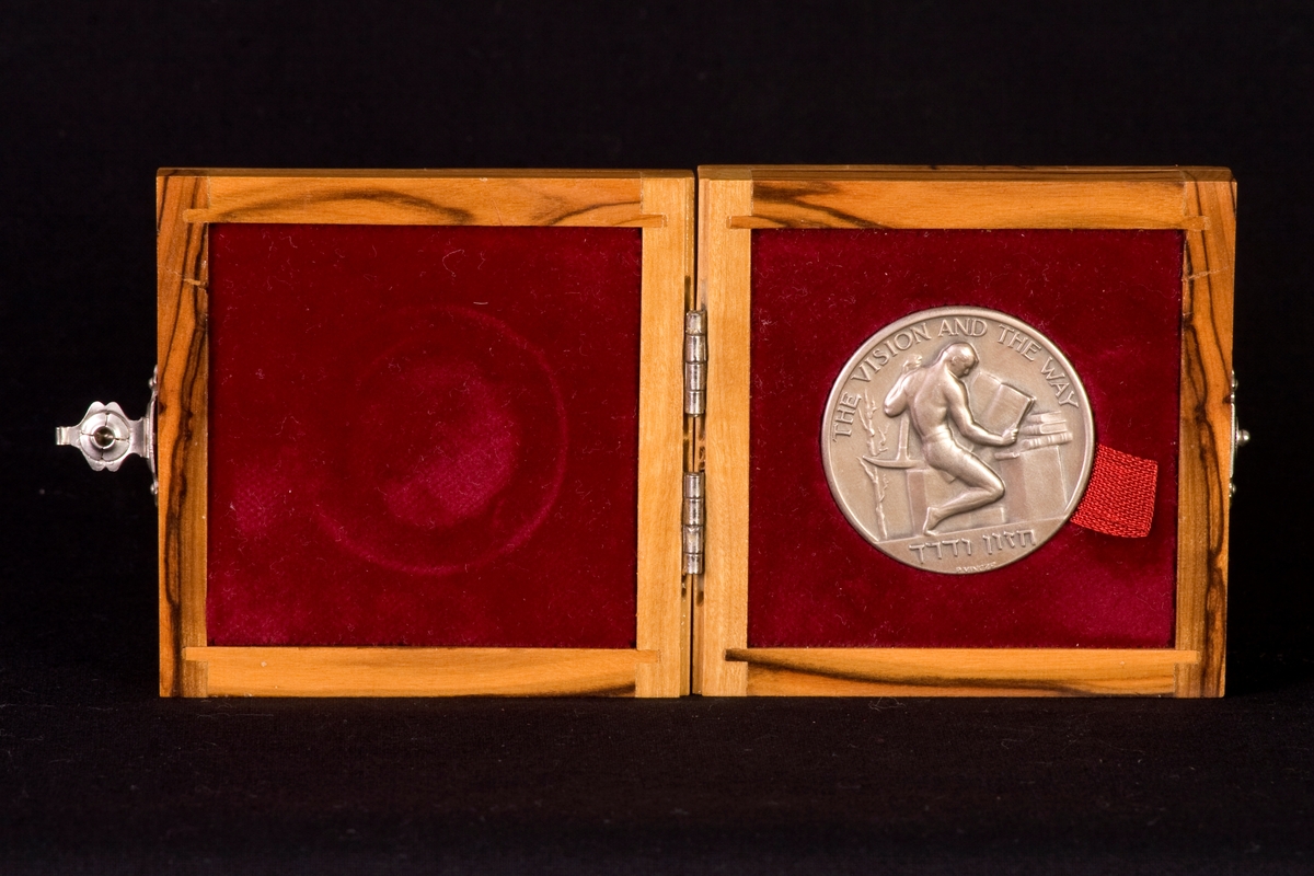 Bronsmedalj med tillhörande träetui, klätt invändigt med röd sammet. Lås på etuiets framsida, tillverkad av metall. Medaljen är dekorerad med text på fram- och baksidan, samt en läsande arbetare på ena sidan och en man i profil (David Den Gurion) på andra sidan.