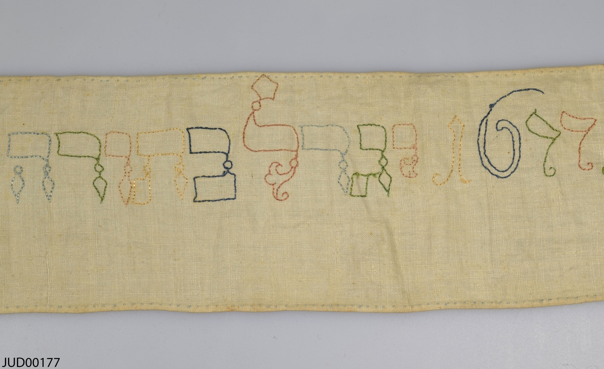 Torabindel (wimpel) tillverkad av linne, som är dekorerad med broderi i olika färger i form av hebreisk text och året 1677.