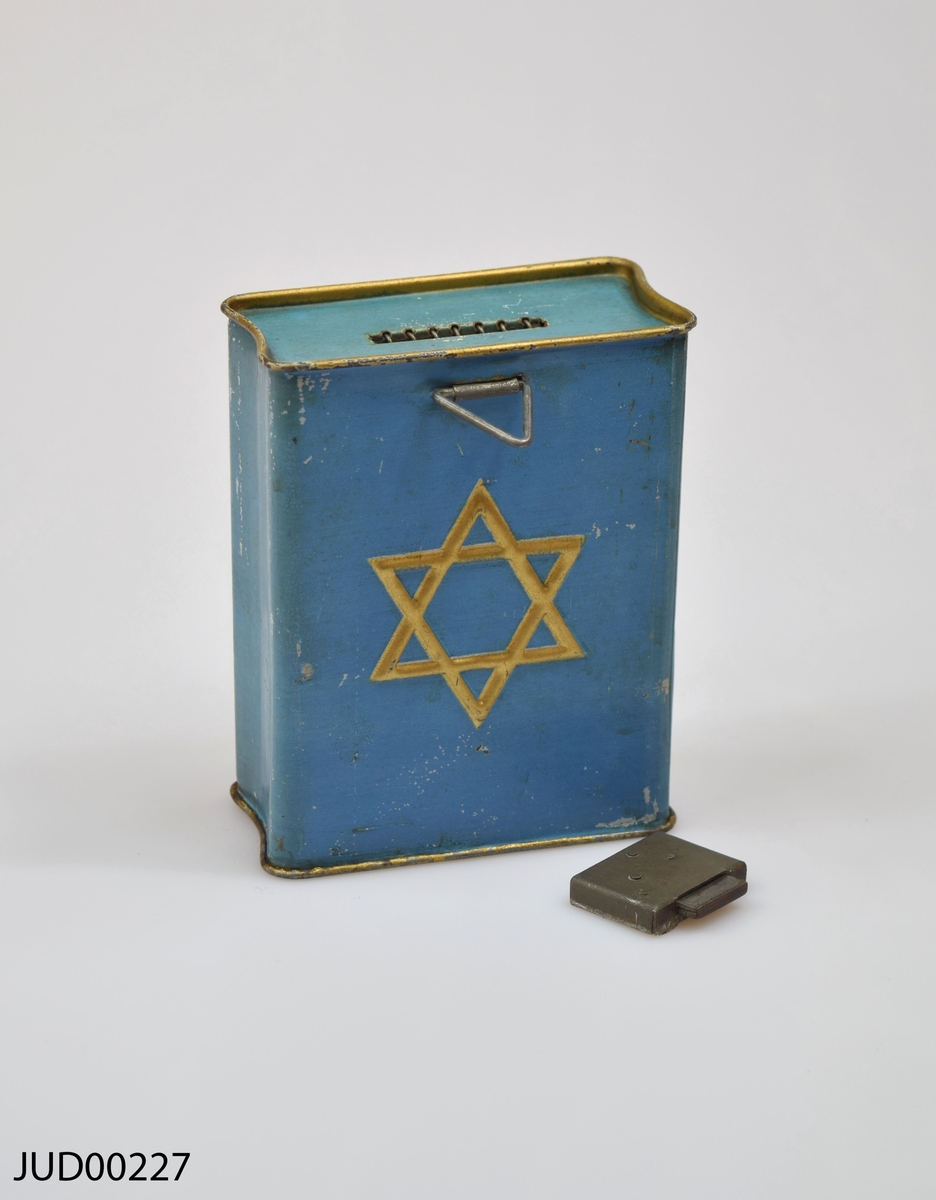 Insamlingsbössa för KKL, tillverkad av plåt. Bössan är målad blå, med en gul Davids-stjärna på framsidan samt hebreisk text. "J.F".