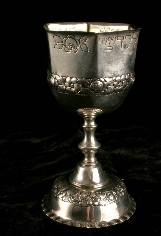 Silverbägare med rund fot och sexkantig cupa. Dekorerad med blommor i relief och ristade hebreiska bokstäver.