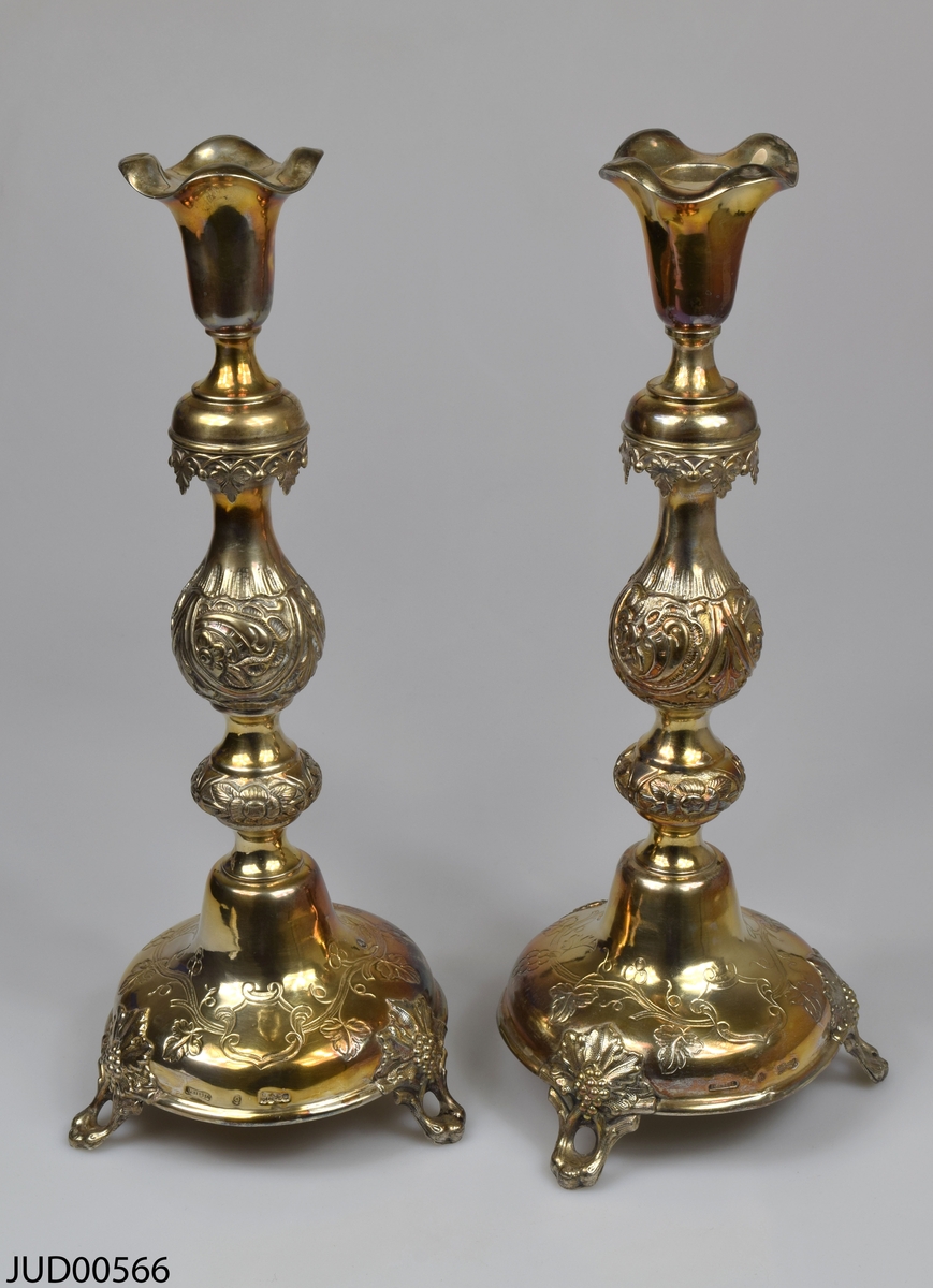 Ett par silverljusstakar med bullig form ståendes på tre fötter. Dekorerad med blommor i relief. Troligtvis ryska stämplar, i form av dubbelörn, 1887, A. Riedel, OC. 84.