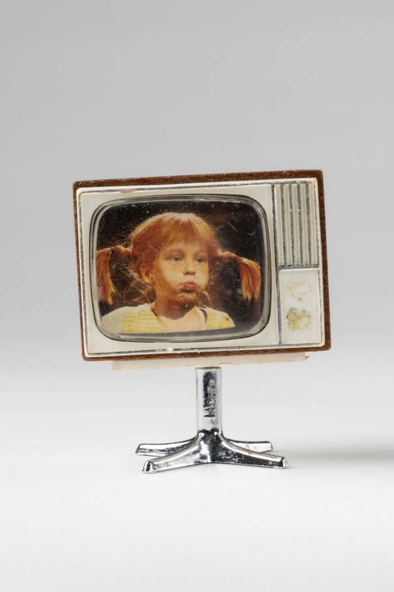 TV till dockskåp, av brunt trä, vit plast på fram- och baksida. Fot av silverfärgad plast. I rutan färgfoto av Pippi Långstrump.