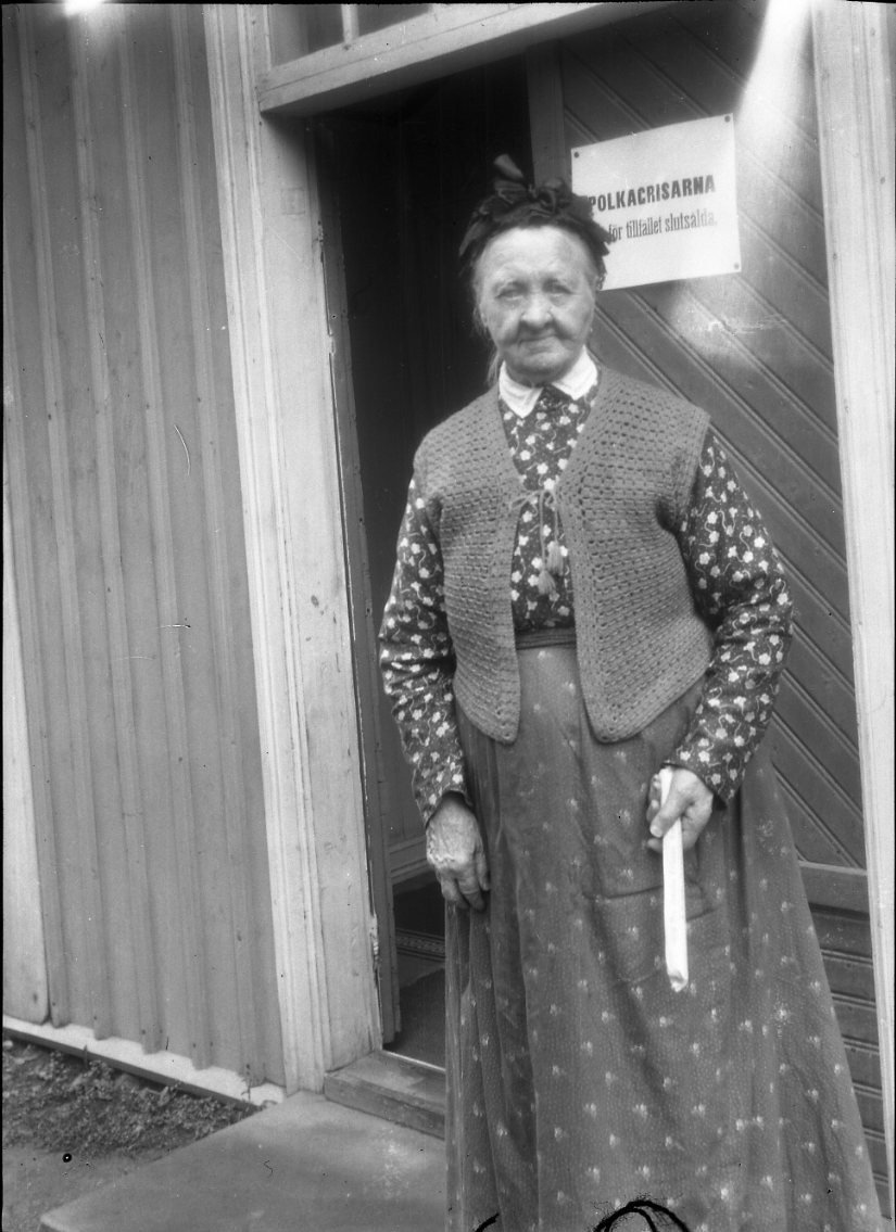 Sockerbagaren Amalia Erikson står i en dörröppning med en polkagris i vänster hand. Hon bär en