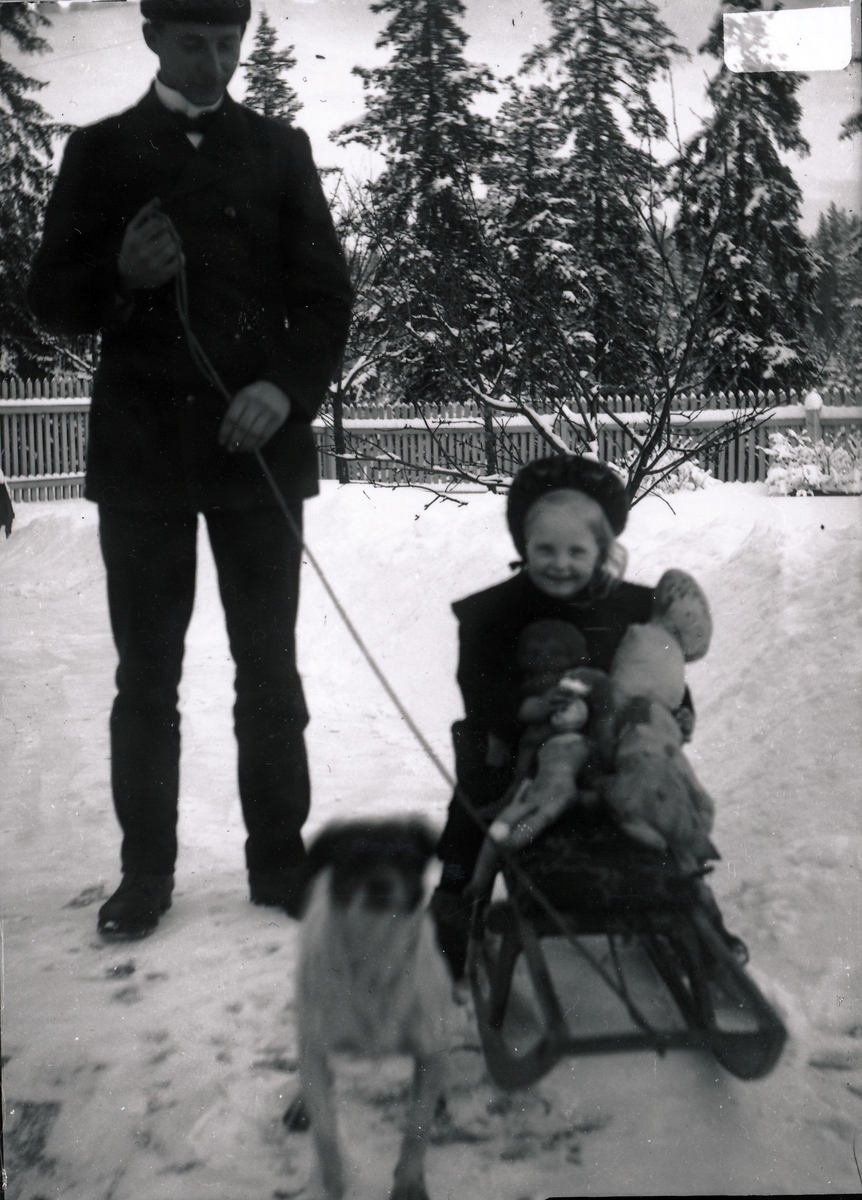 Bestyrer Wilhelm Rydgren og datteren hans Inger Elisabeth, kalt Lisbeth, på kjelke i hagen tilhørende bestyrervillaen på Kistefoss.  Lisbeth har dukker og bamser i armene, og en liten hund står i forgrunnen.