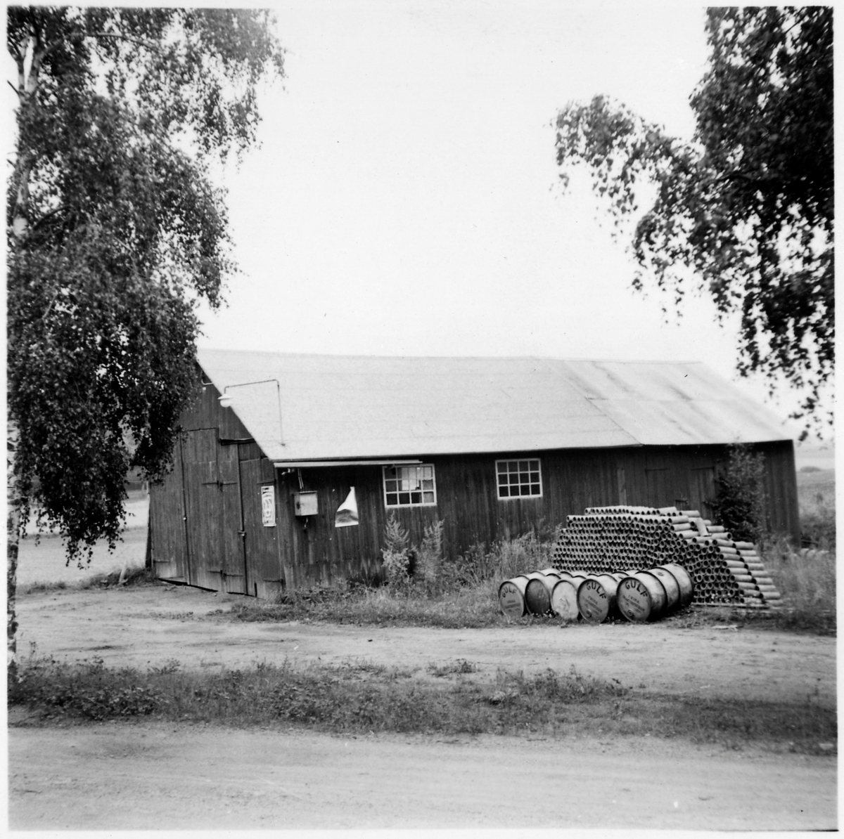 Vägstation E10, Östra Husby, filial Kummelby. Garagebyggnad med port på gaveln. Upplag av täckdikningsrör, muffrör, bränslefat.