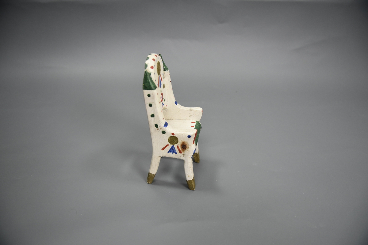 Stol til dokkemøblement i fem deler. Denne stolen er litt mindre enn de tre andre. Møblene er hvitmalte med dekor i ulike farger. 
Møblementet er fra givers tidlige barndom, muligens 1928-1930. Møblene var til hennes første dokkestue, bygget av far av en appelsinkasse. 