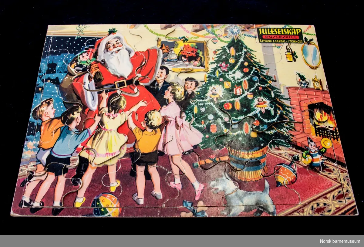 Puslespillet er et julebilde, hvor nissen kommer med gaven til et juleselskap med mange barn som er samlet i en koselig julestue. 