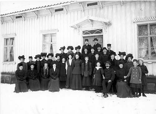 En grupp på 33 män, kvinnor och barn står uppställda i vinterkläder på trappan till Uppgränna Mattsagård. Det är vinter och snö på marken.