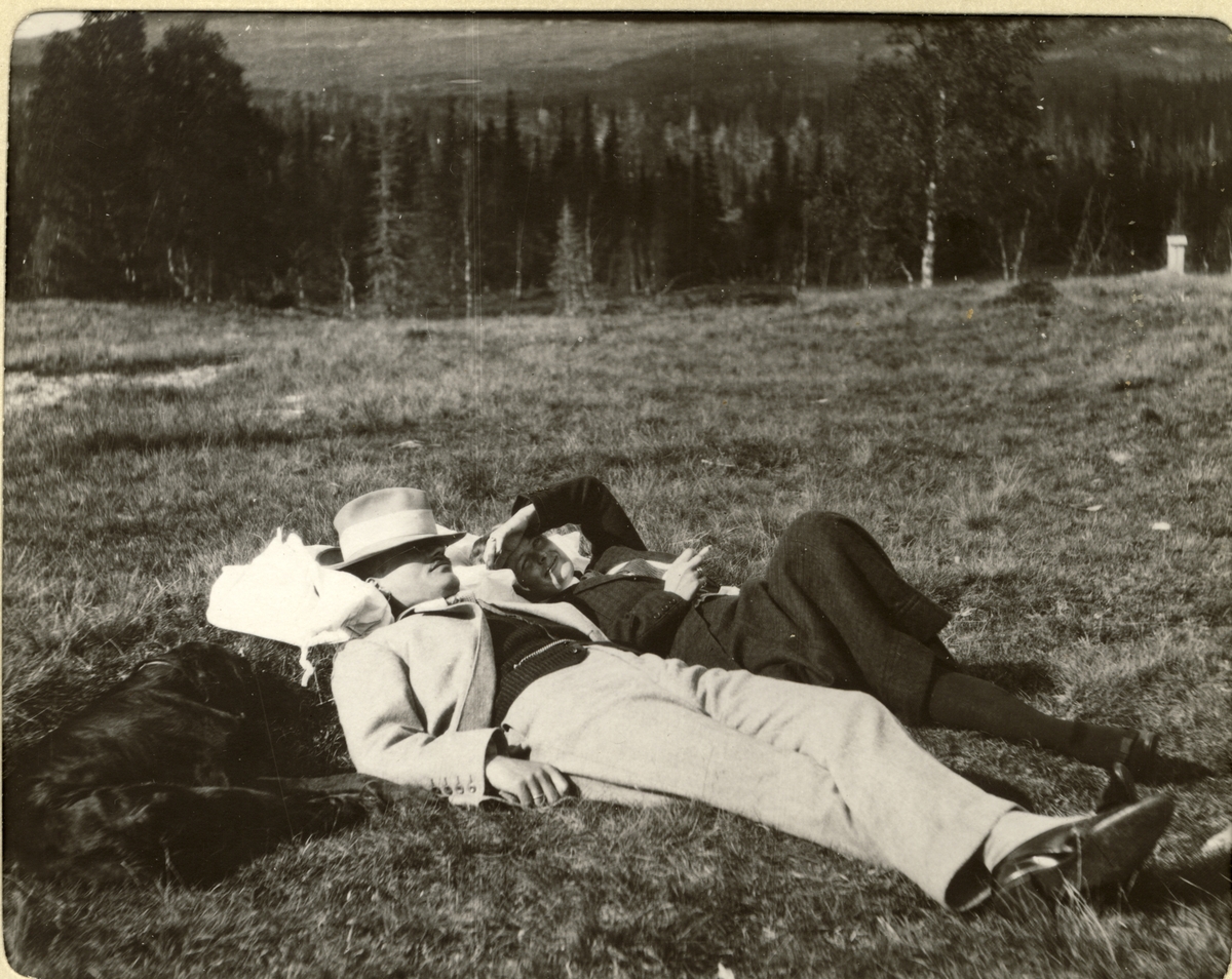 Et par, trolig Gina og Herbert Plathe, og en hund ligger på en åpen slette og nyter søndagsfreden under rypejakt i Meråker, Nord-Trøndelag. Fotografert 1910.