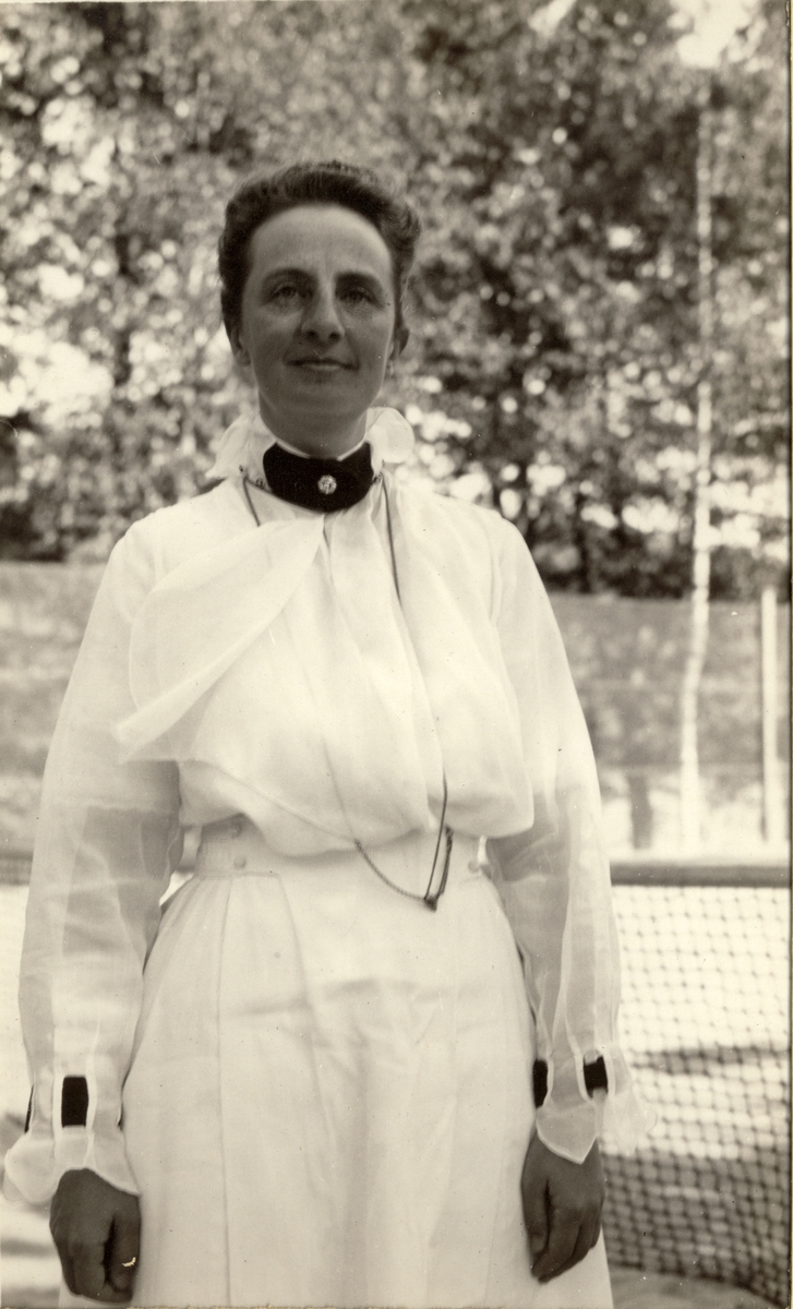 Nini Egeberg, antagelig stående på en tennisbane. Nettet skimtes i bakgrunnen. Fotografert 1920.