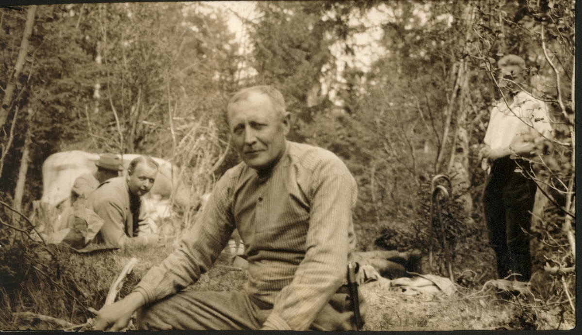 Helge O. Furuseth sitter i en åpning i skogen. Tre ukjente menn i bakgrunnen. Fotografert 1923.