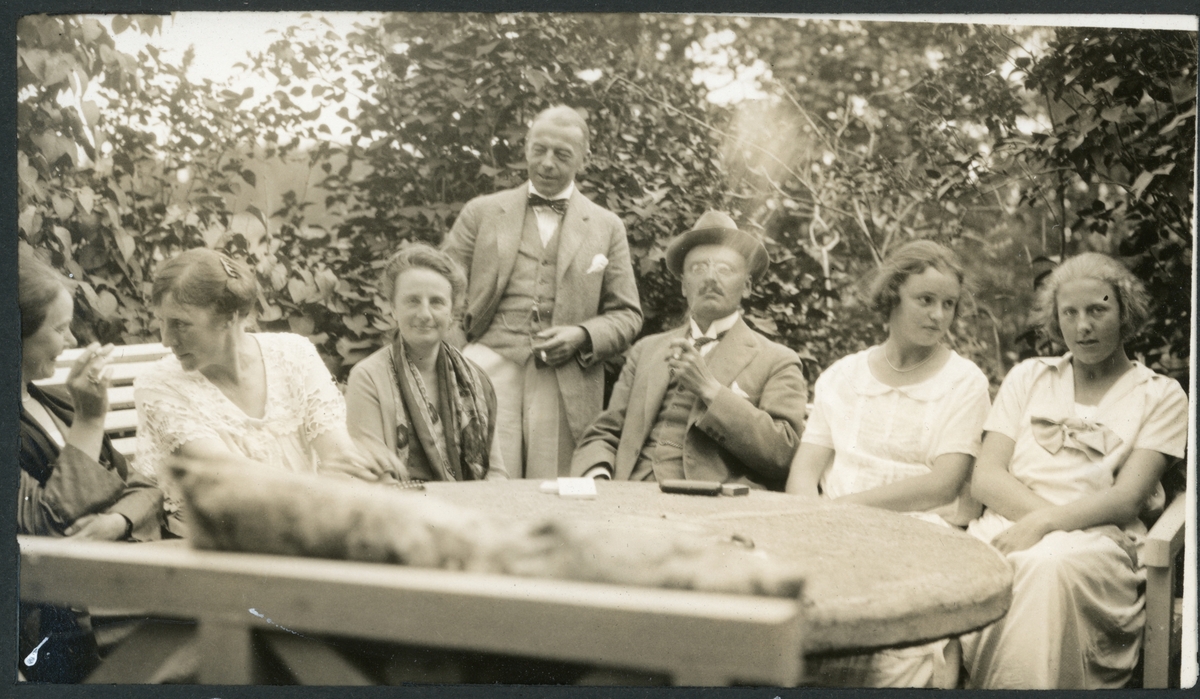 Syv personer samlet rundt et hagebord på Brekke gård, i Oslo(?). Fra venstre ses Mabel Butenschøn, Nini Blom, Nini Egeberg, Christian Blom, Barthold Butenschøn, Lille Mabel Butenschøn(?) og Aimé Blom. Fotografert august 1924.