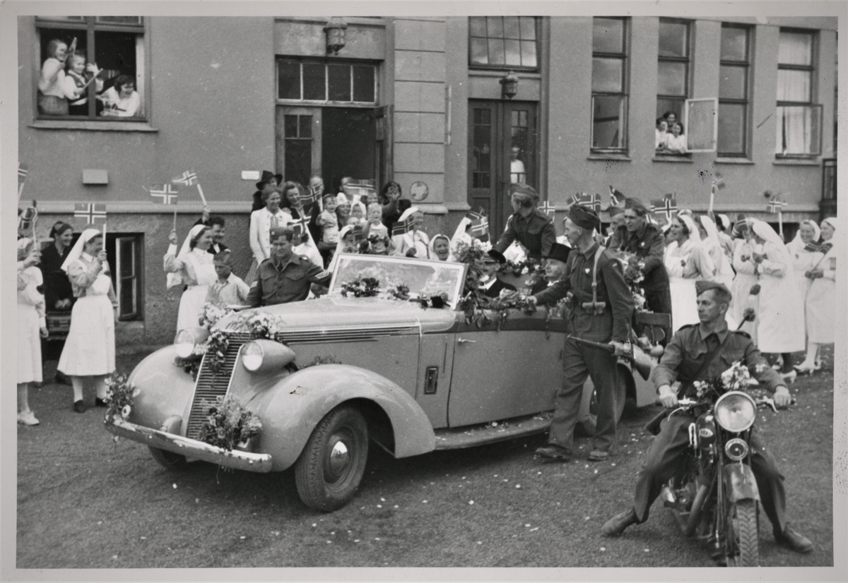 Den 22. august 1945 besøker kong Haakon VII Haugesund. Her er kongen i den pyntede "kongebilen" utenfor medisinsk avdeling på Haugesund Sykehus. Sykepleiere med norske flagg har kommet ut til "kongebilen". Menn fra politistyrkene er på vakt for kongen.