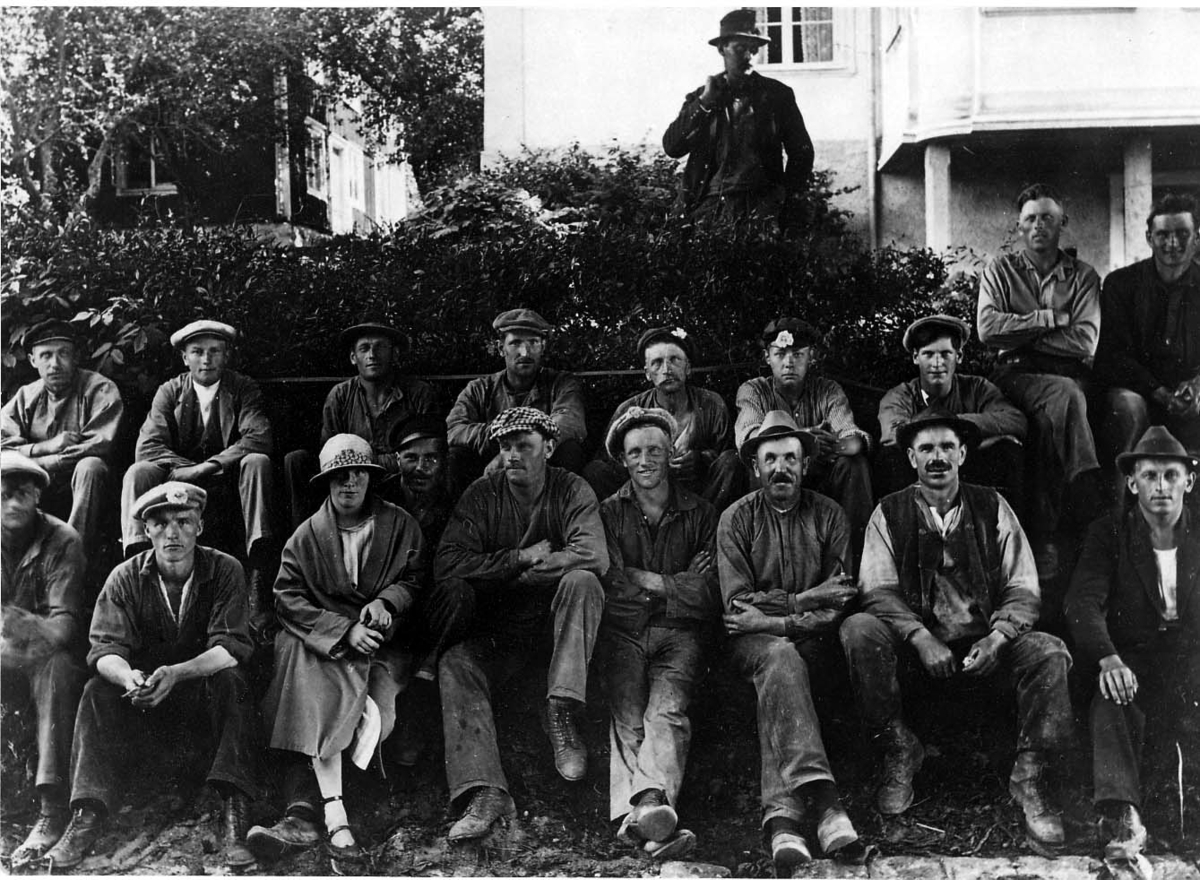 Gruppbild med ett 10-tal män i arbetskläder och en kvinna som sitter på en grässlänt till en fastighet.