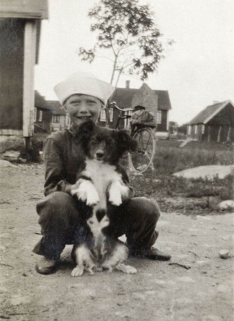 En liten pojke (?) visar fram en hundvalp. I bakgrunden skymtar en cykel och några bostadshus m.m. 
Under fotot text: "Hällevik, 1929".