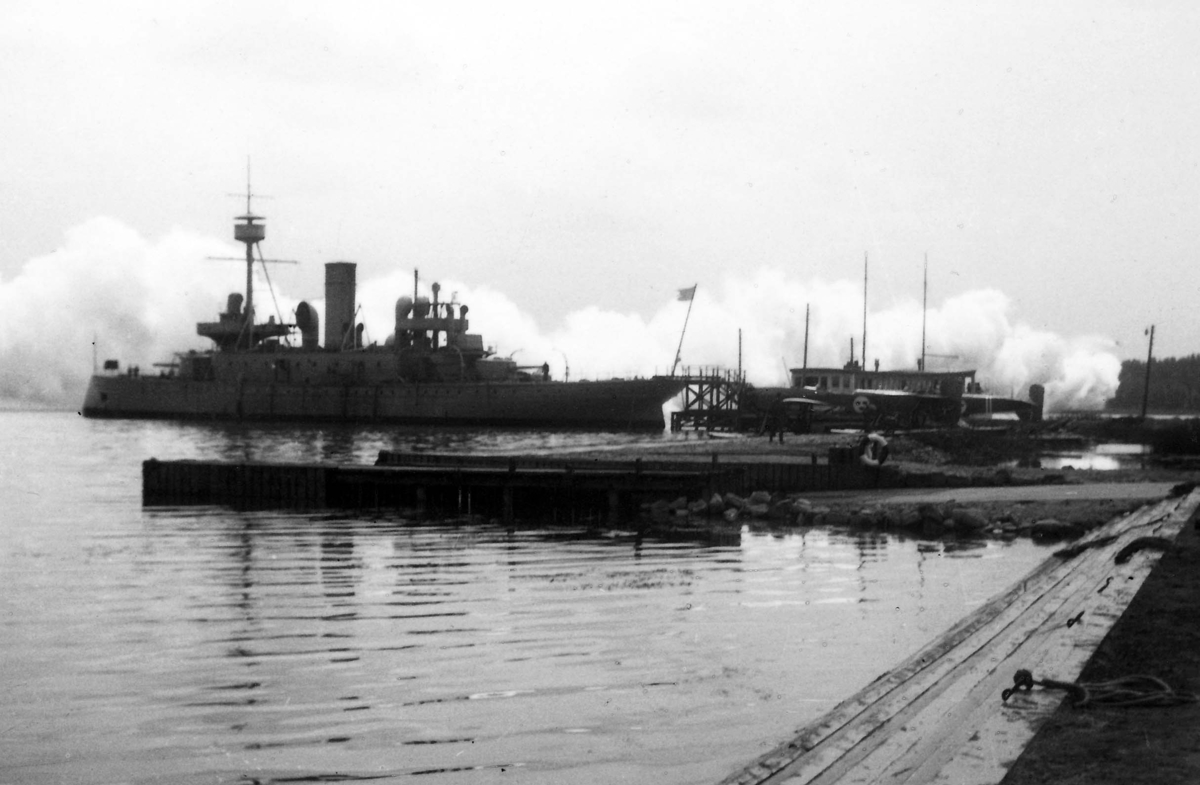 Vy över hamnområde med marinfartyget Dristigheten vid kaj och ett militärt sjöflygplan. I bakgrunden syns ett stort moln över vattenytan. 1930-tal.