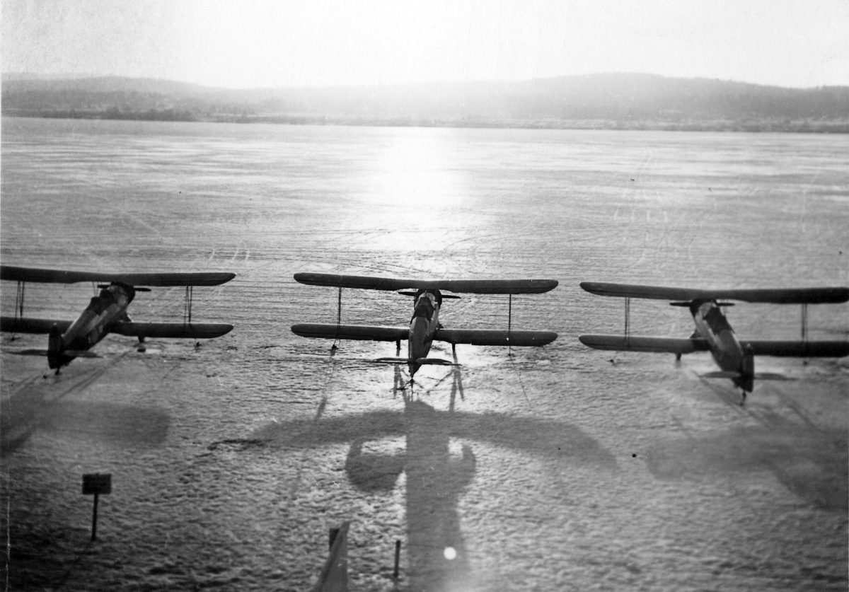 Översiktsbild av tre flygplan Sk 12 ståendes på en sjö, vintertid. Vy bakifrån, med motljus och berg i bakgrunden. Cirka 1936-1939.