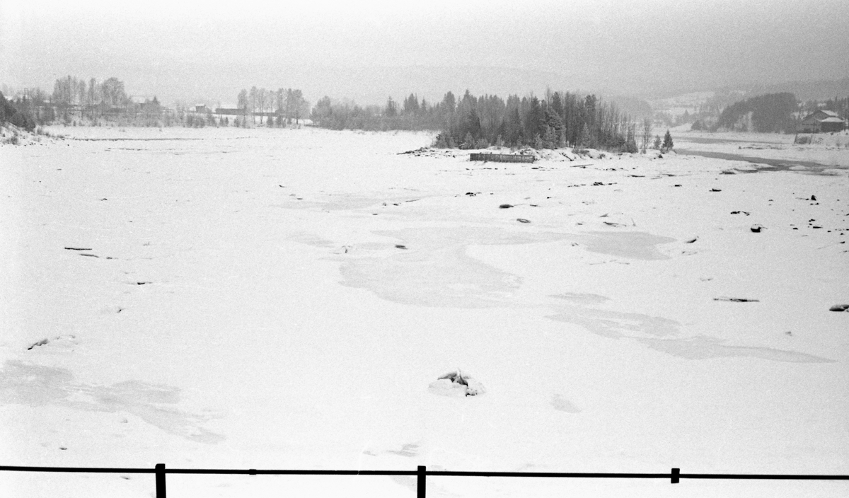 Skjefstadfossen i Glomma, i Heradsbygda i Elverum i Hedmark. Fotografiet ble tatt vinteren 1960, fra en forholdsvis høy plattform ved kraftverksdammens østre landfeste. På dette tidspunktet var vannføringa minimal, og fossen var dekket av is og snø. Bare en del steinblokker stakk opp. På motstrøms side av Tjuvholmen, som delte elveløpet og fossen i to, skimter vi en skjerm, ei steinfylt tømmerkiste, som skulle forebygge at fløtingstømmeret, når våren kom, drev inn på holmen og måtte bæres tilbake mot elveløpet igjen når vannstanden sank.