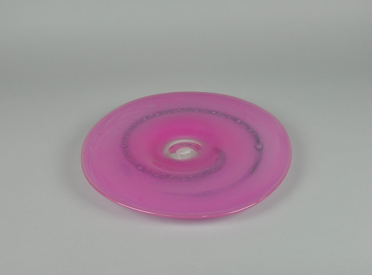 Munnblåst fat av glass. Rund form, med sokkel som er utledet fra resten av fatet. Glasset er farget rosa med en lilla spiral, og klart i midten.