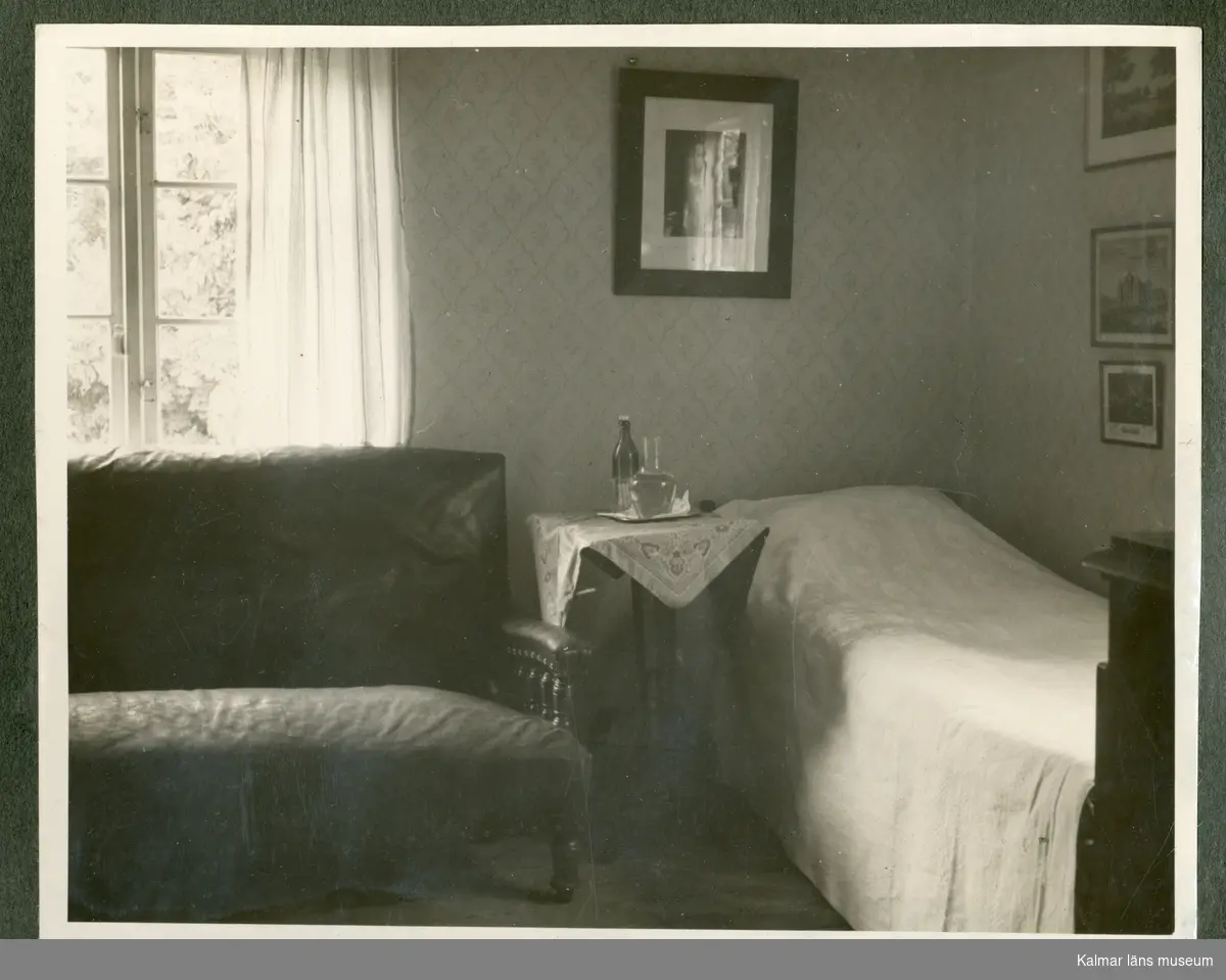 Svart-vitt fotografi, interiörbild. Framför ett fönster till vänster står en soffa. Till höger står en säng. Ovanför sängen hänger tavlor på väggarna. Bredvid sängen står ett bord. Troligtvis interiör från Waldemar Swahns sommarbostad på Stångudden, Ekenäs.