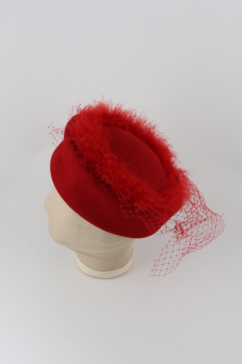 Rød sirkulær hatt i stiv filt. Bord av syntetiske røde fjær rundt hattens topp. Rød netting festet under fjærene henger ned på sidene og ved bakparten.