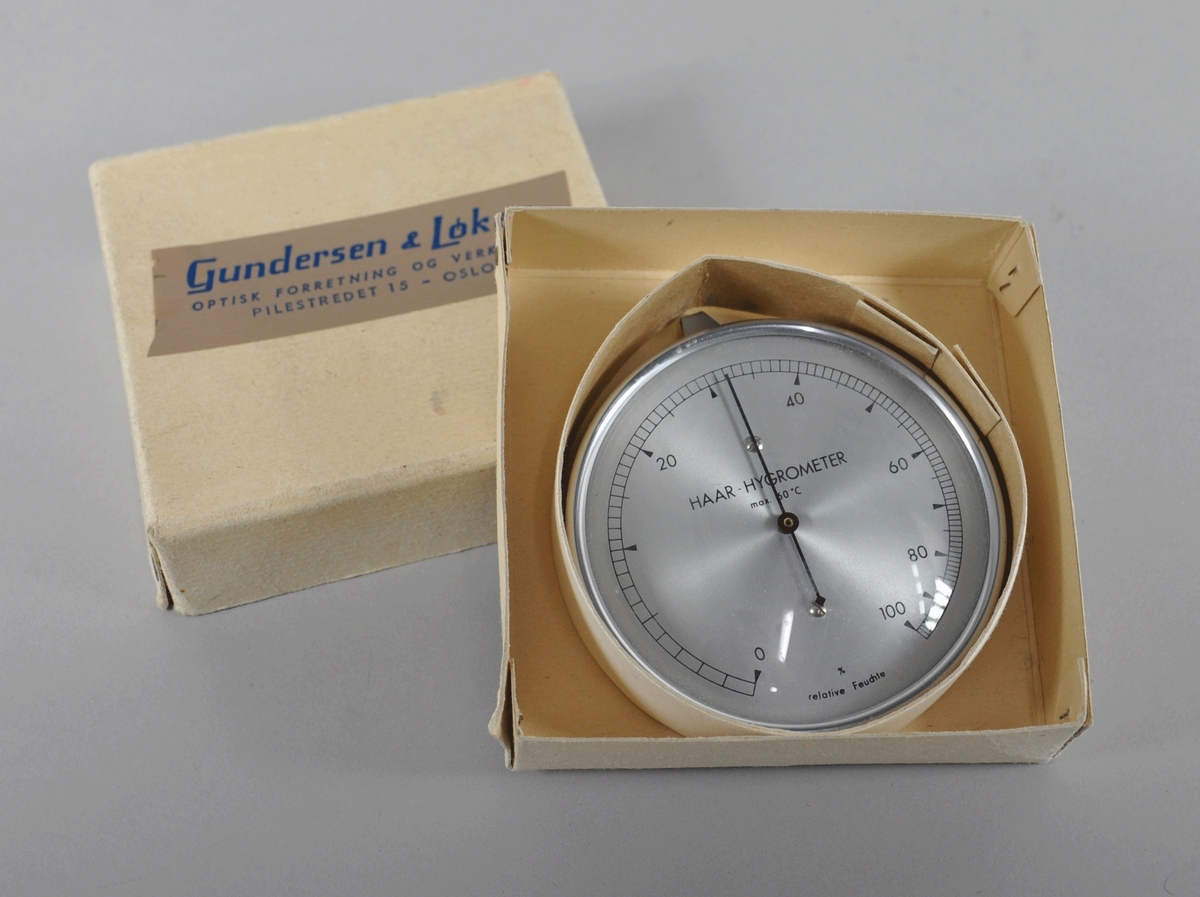 Rundt hygrometer med to visere, som måler fuktighet. Hygrometeret ligger i original emballasje.
