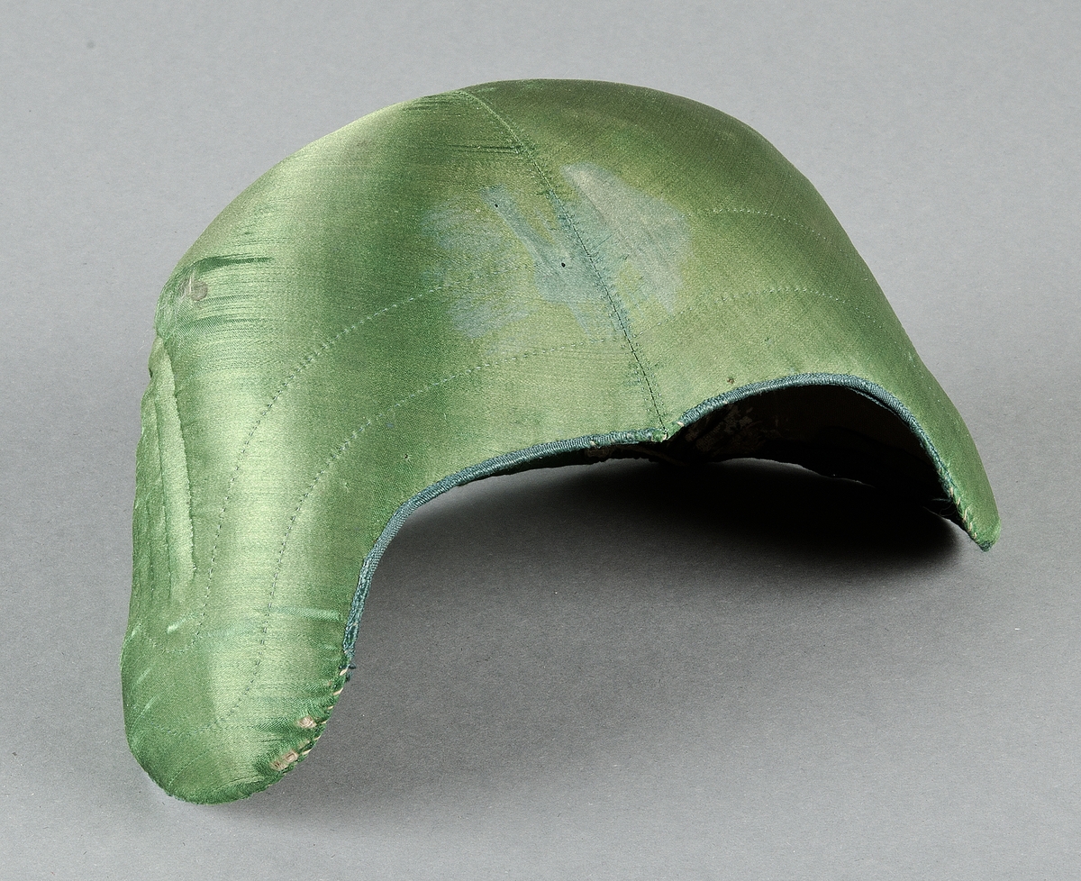 Bindmössa av grönt siden. Fodrad med olivgrön lärft. Bredd över hjässan från sida till sida 35 cm. Längd över hjässan 20,5 cm.