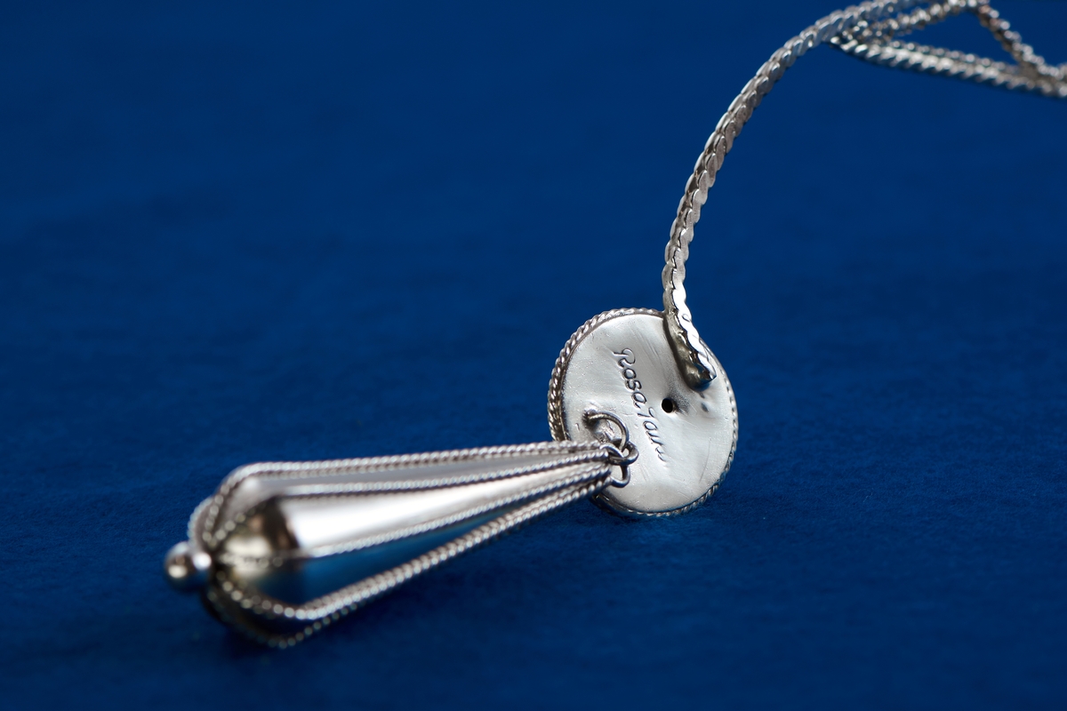 Silversmycke i filigranteknik av Rosa Taikon. Tunga, droppformade örhängen är vanliga smycken hos flera romska grupper. De kan vara utförda i såväl solid som genombruten form. Ofta bärs tyngden upp av en tunn kedja som läggs runt örat för att avlasta örsnibben. I det här huvudsmycket har Taikon valt att istället låta tyngen bäras av huvudet genom att fästa hängena i ett diadem av filigran.