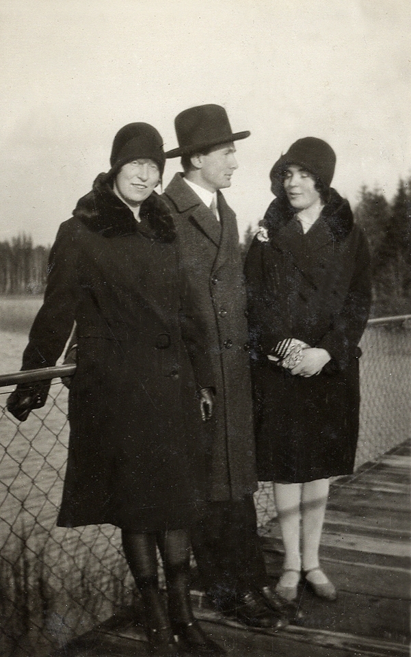 Två kvinnor i hatt och kappa står tillsammans med en man på en stängslad brygga (?) (spång?). 
Text under fotot: "På promenad, 1930".