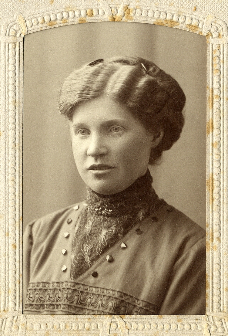 En kvinna i mörk klänning med stenkolsknappar, mörk spetsisättning och hög krage. 
Vid kragen syns en brosch. 
Bröstbild, halvprofil.
