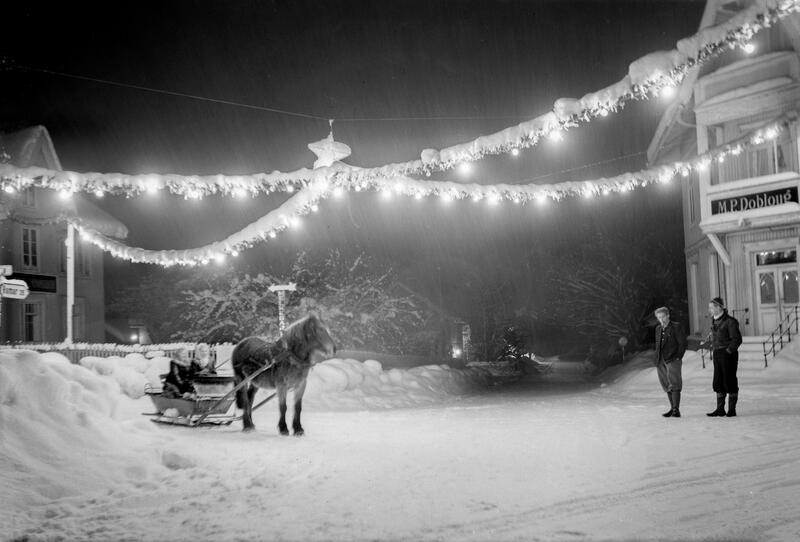 Svart-hvitt bilde av to lyslenker som henger i kryss over snødekket gate, hest med slede til venstre i bildet og to mennesker foran et hus til høyre. (Foto/Photo)