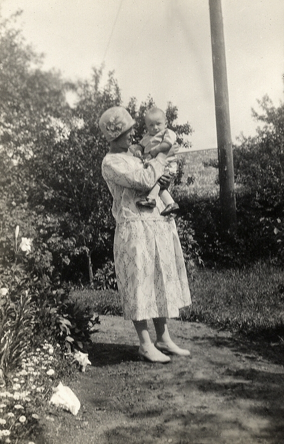 En kvinna i hatt och klänning håller en baby i famnen, i en trädgård.
