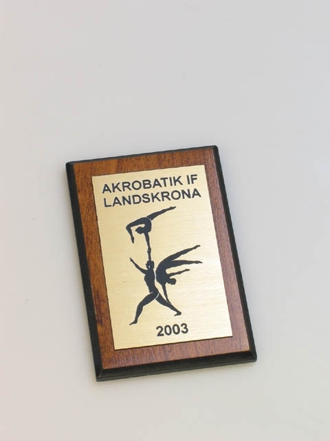 Plakett med texten "AKROBATIK IF LANDSKRONA 2003". Akrobatik IFs logotyp i mitten. Plaketten är av plast med trämålning och guldfärgad metall.
