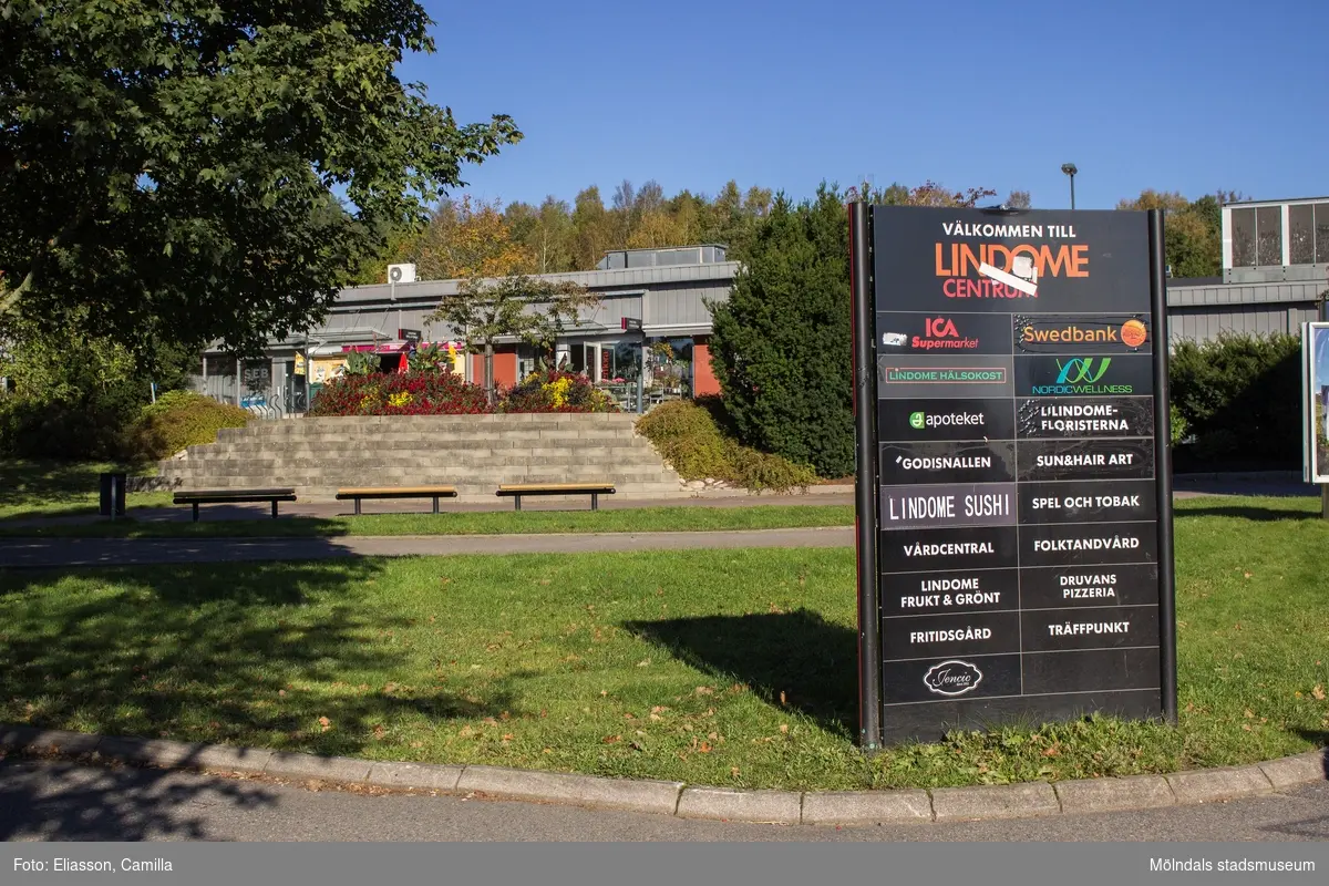Liten park i Lindome centrum den 4 oktober 2016. I bakgrunden ses Lindome Spel & Tobak samt Lindomefloristerna i huset Almåsgången 2. I parken finns sittplatser samt en stor skylt som anger vilka affärsrörelser med mera som finns i centrum. Vy mot norr.