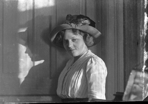 Bröstbild av en ung kvinna i ljust randigt livstycke och stor hatt. Hon står lite i profil mot en vägg. Hennes skugga faller till vänster på bilden.