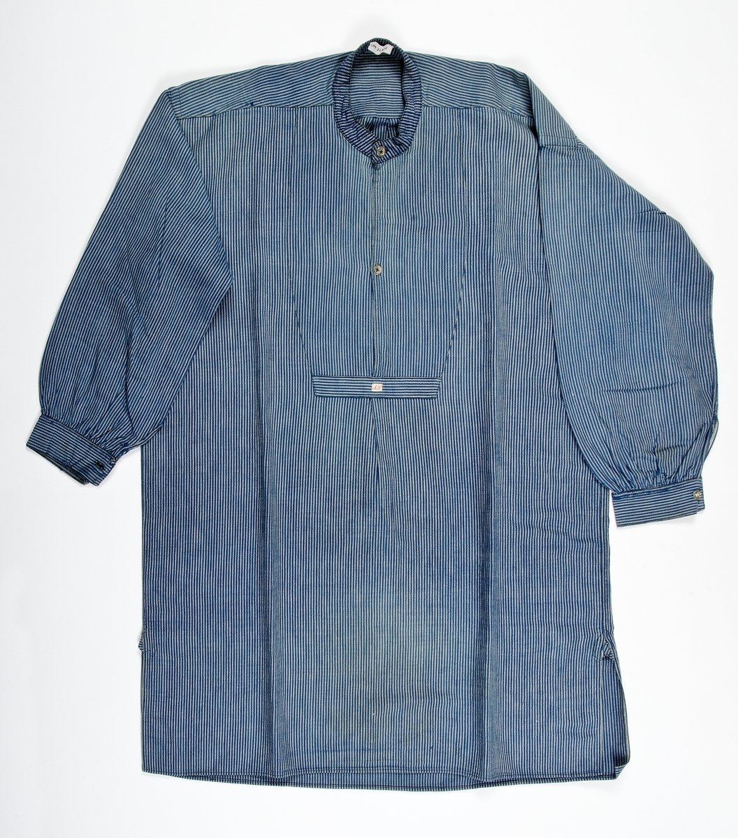 Skjorta av blåvit-randig bomullskypert. Rak med axelok och med sprund fram och dubbelt tyg i bröstpartiet avslutat med tvärslå, två metallknappar och ståkrage. Lång ärm med ärmspjäll och manschett med knapp.
