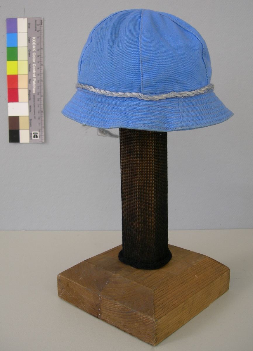 Rundkullig hatt sydd av 6 kilar med litet brätte. Ljusblå bomullskypert, dekorerad med tvinnad konstfibersnodd i ljusblått och vitt. Fodrad med gulvitt bomullstyg.