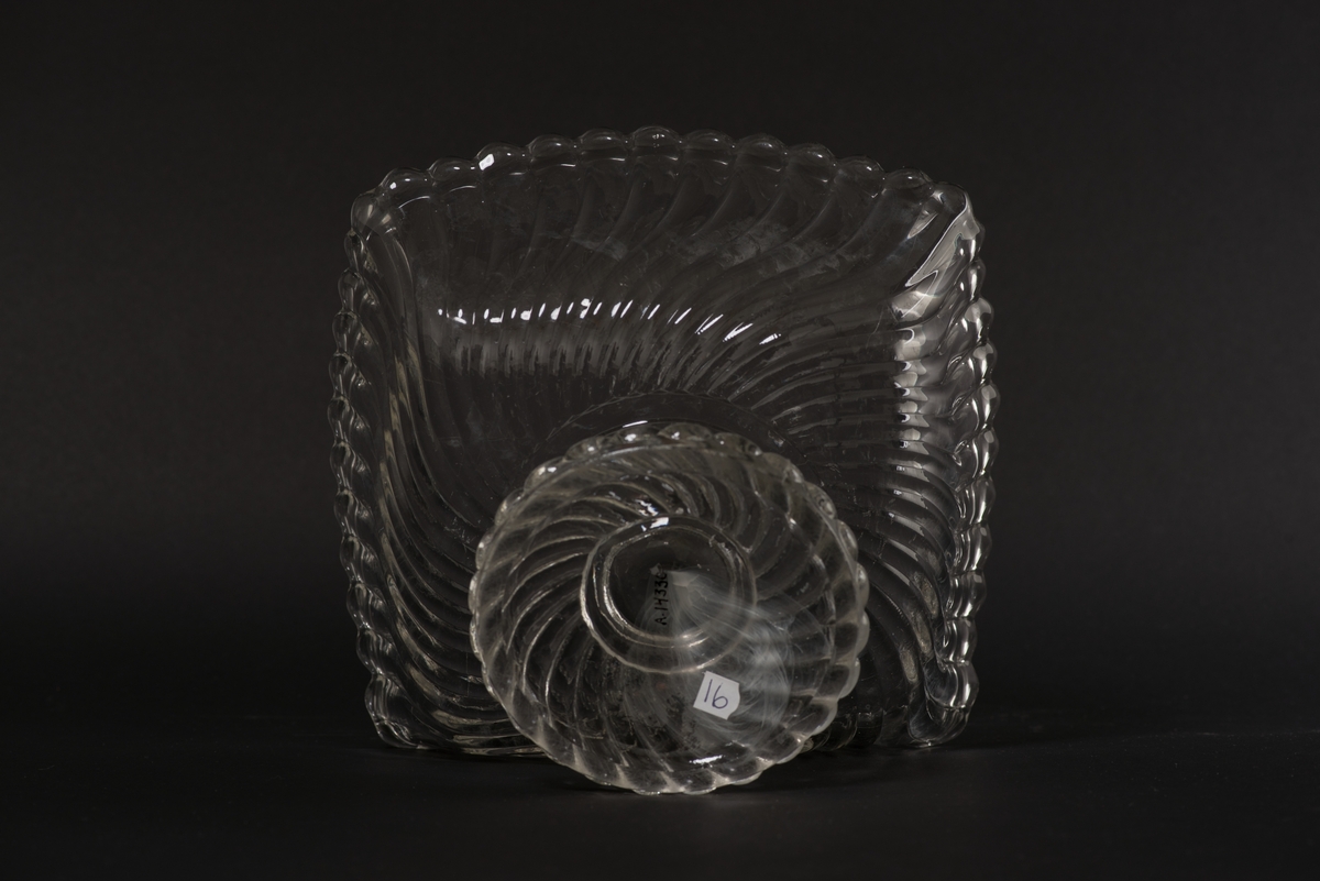 Fyrsidig skål på fot av ofärgat pressglas. Pressad yttäckande dekor av spiralvridna åsar som avslutas i en vågformad kant.