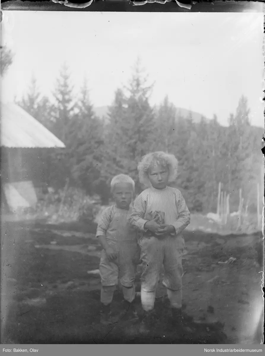 To barn stående oppstilt utendørs. Deler av en bygning i bakgrunnen.