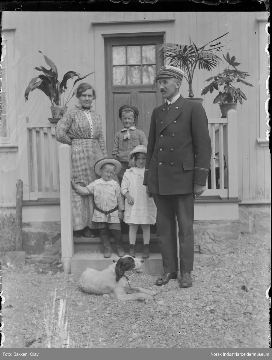 Ramilien Rasmussen stående utenfor stasjonsmesterboligen på Gransherad stasjon. Stasjonsmester Sigurd Rasmussen i uniform. Familiens hund "Sjakk" liggende på bakken.