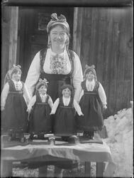 Dame i bunad stående bak bord med fire dukker kledd i bunad.