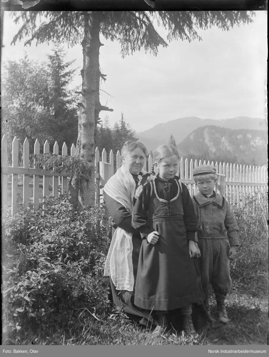 Kvinne sittende i hage med to barn stående foran seg. Kvinnen har sjal rundt skuldrene.