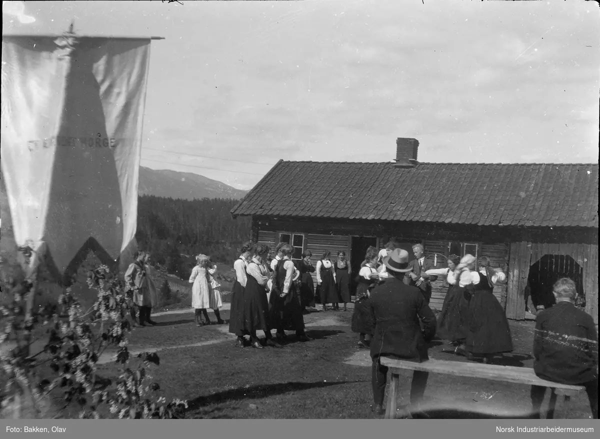 Menn, kvinner og barn i sosial sammenkomst med dans utenfor bygning. En fane med påskriften "Et xxxxxx Norge".